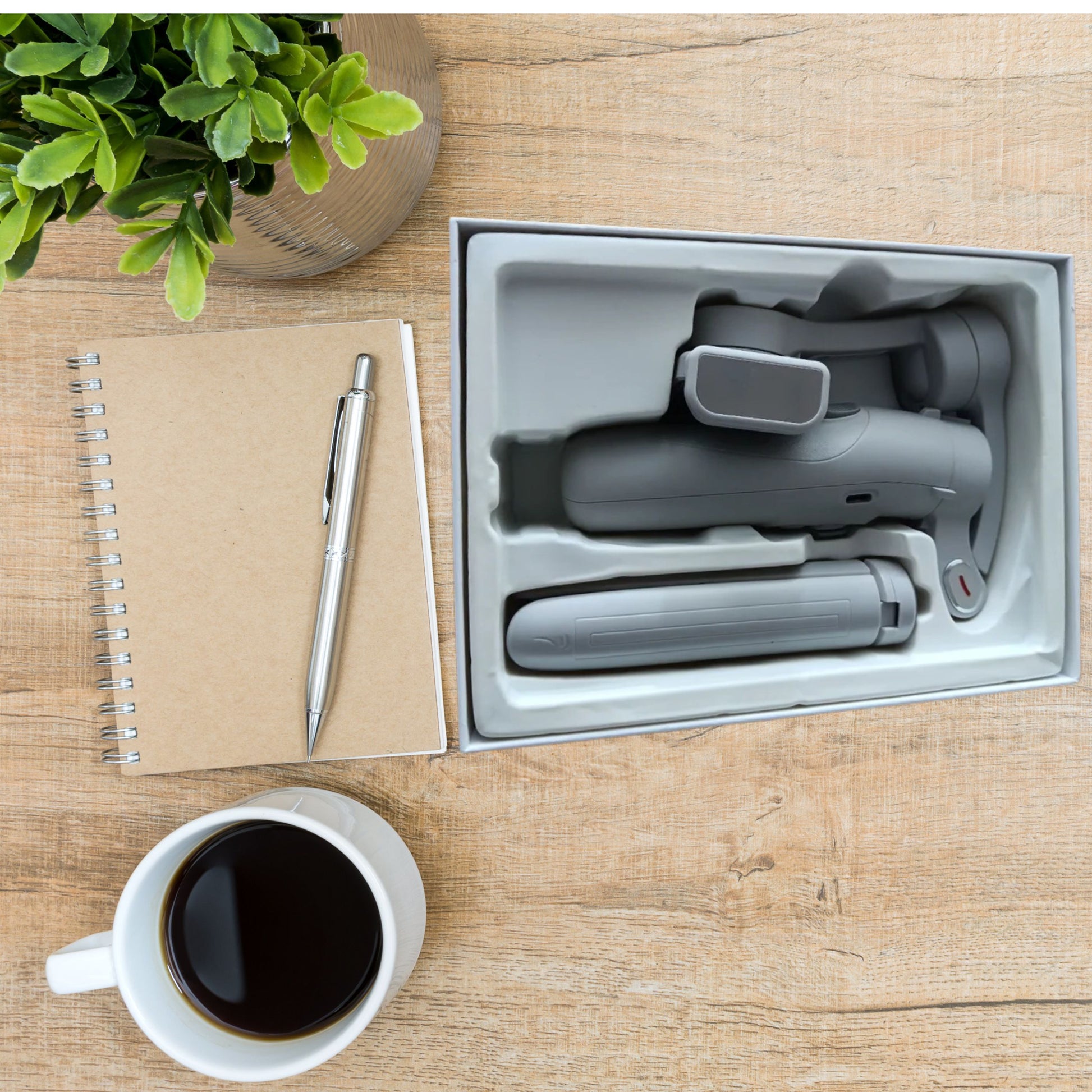 Gimbal estabilizador para móvil 3Ejes en caja, con cuaderno y café en la mesa.